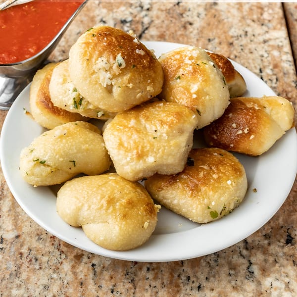 A plate of golden-brown garlic knots with parmesan cheese and parsley, served with a side of marinara sauce.